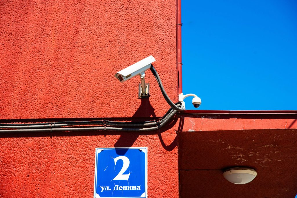 A vivid red wall featuring surveillance cameras and a number sign under a clear blue sky.