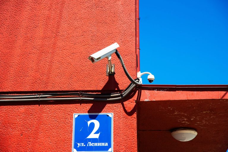 A vivid red wall featuring surveillance cameras and a number sign under a clear blue sky.